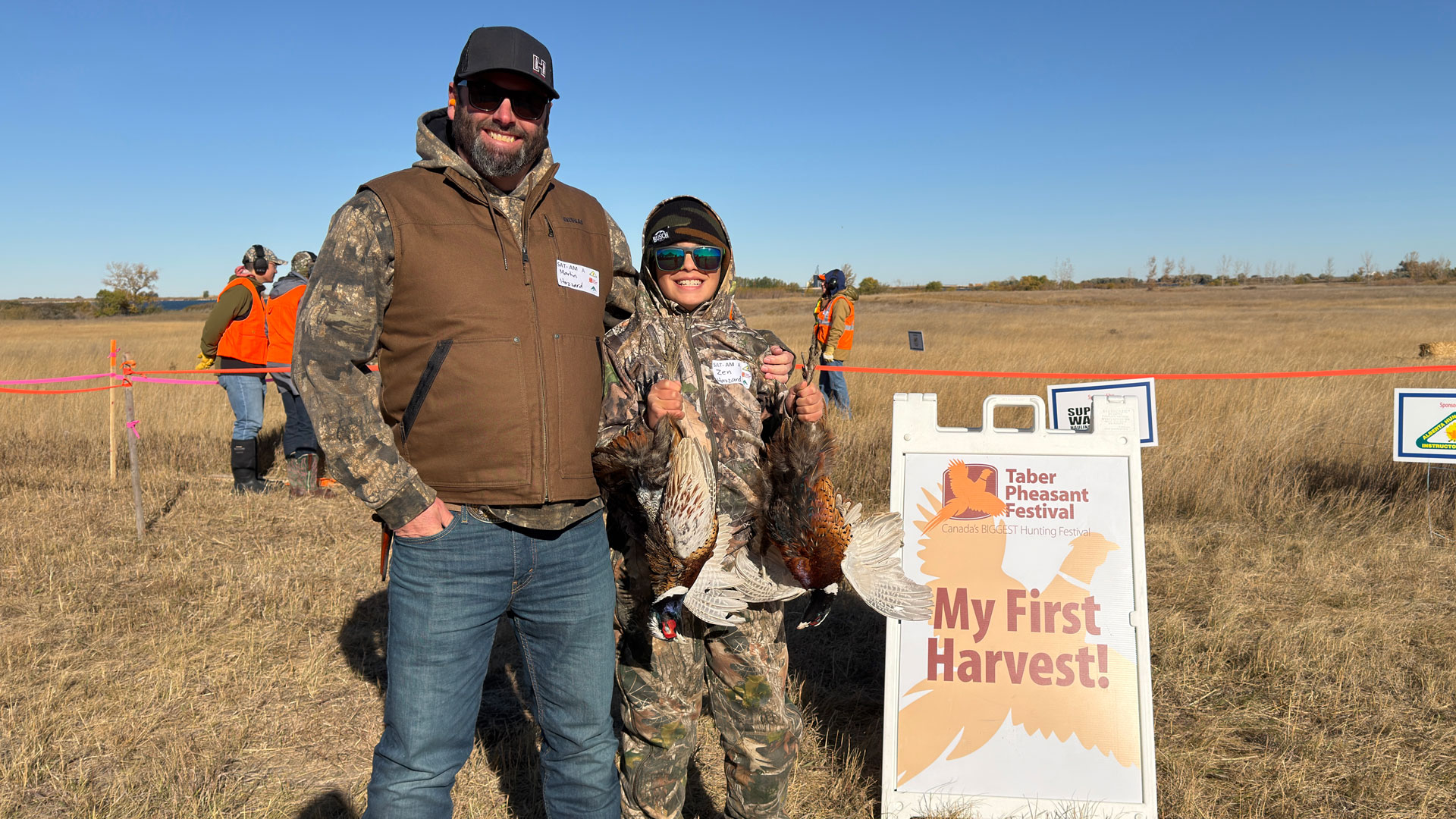 Kid holds up 2 dead pheasants in front of a sign that says My first Harvest