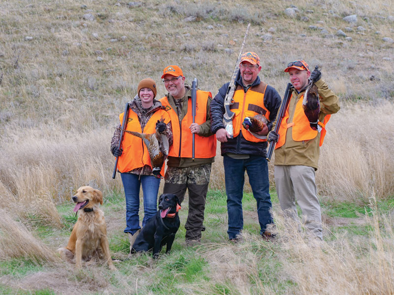 4 hunters stand with their rifles and dogs in a field